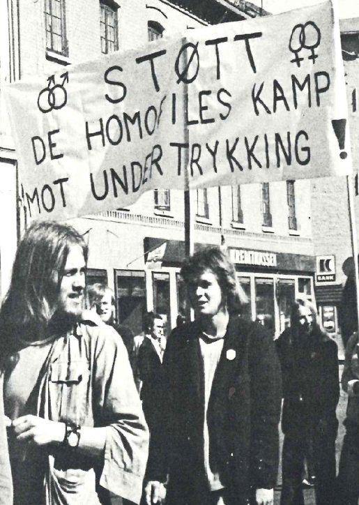 Fra pride-markering i Oslo i 1974. Svart/hvitt bilde med folk som holder banner hvor det står "støtt de homofiles kamp mot undertrykking"
